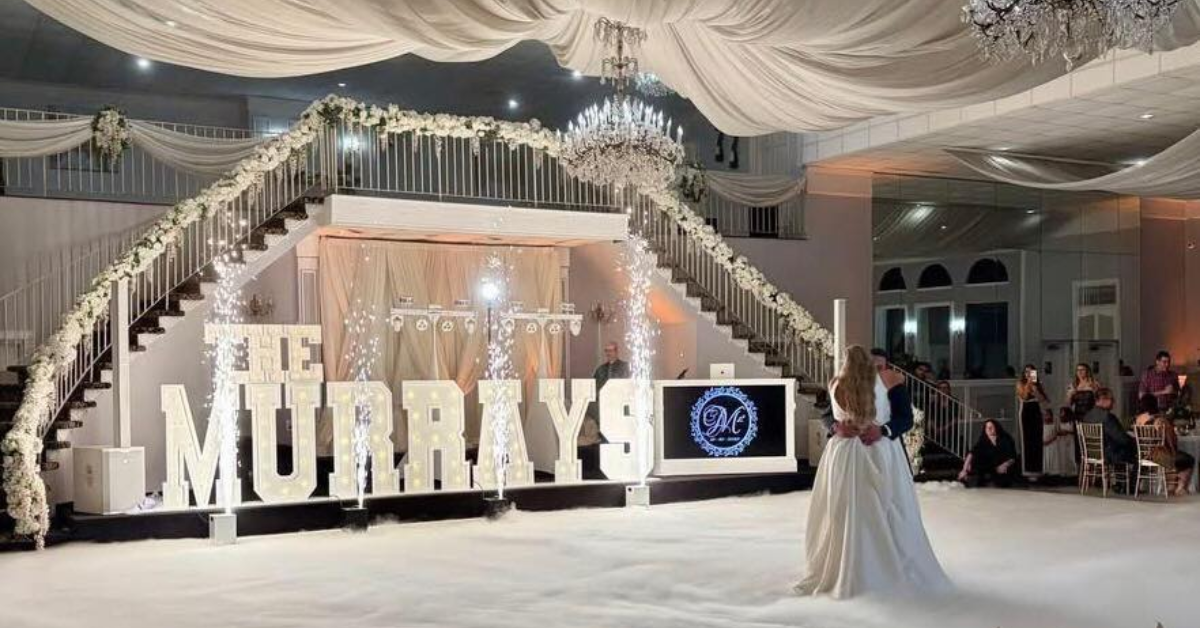 A bride and groom share a first dance in a lavish ballroom with draped ceilings. Large Light-Up Marquee Letters spelling "The Murrays" and floral decor create an elegant atmosphere.