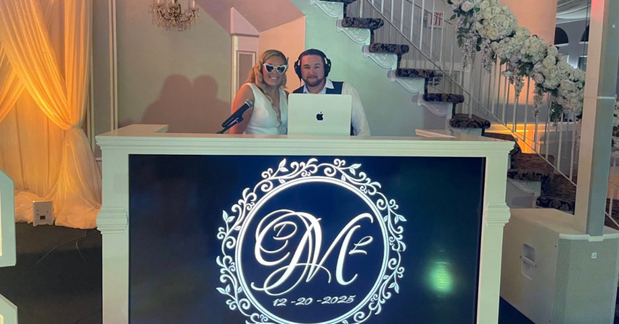 A couple stands behind a DJ booth decorated with floral designs and initials "DM," dated 12-20-2025. An elegant staircase and soft lighting set a celebratory mood.