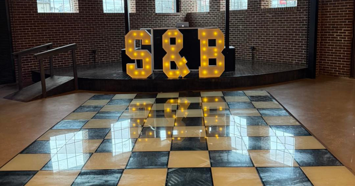 Dimly lit brick-walled room with bright marquee letters "S & B" on a stage, overhead industrial light fixture, and reflective checkered floor.