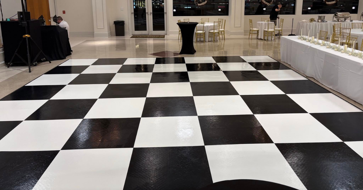 A ballroom with a glossy black and white checkered dance floor, elegant chandeliers, draped ceiling fabric, and tables set with white linens and gold chairs.