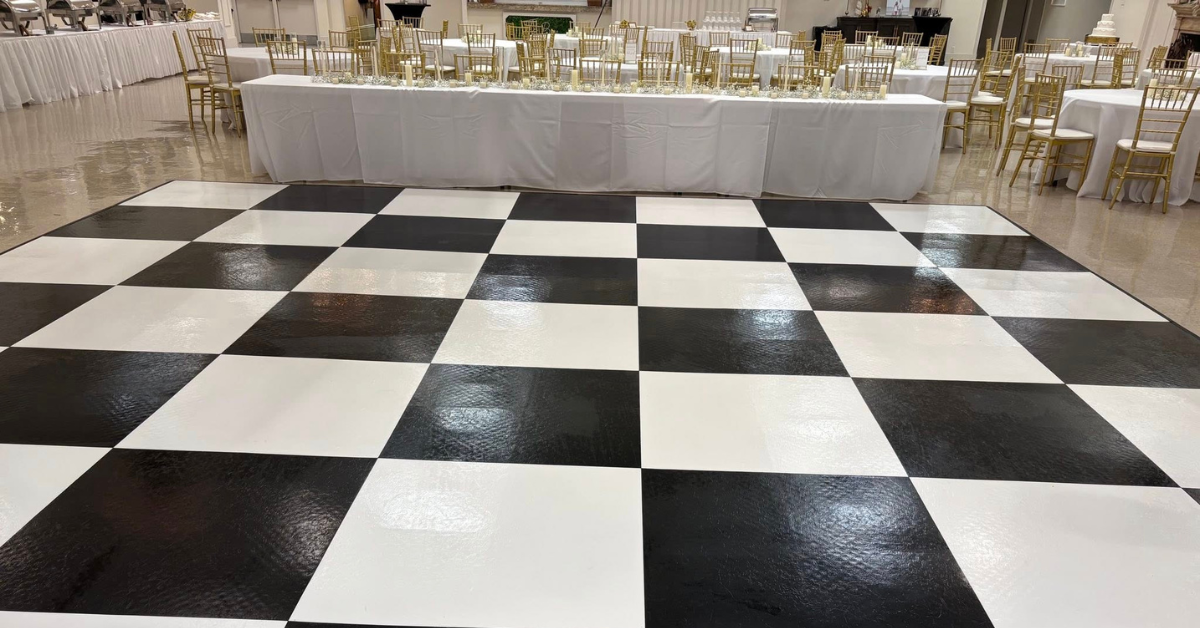 A polished black and white checkered dance floor is in the foreground, with a long table and gold chairs draped in white cloth behind it, suggesting a formal event setup.