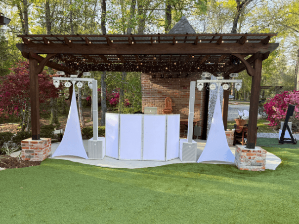 Outdoor DJ setup under a wooden pergola surrounded by spring foliage. The area is decorated with white speakers and lights, creating a festive ambiance.