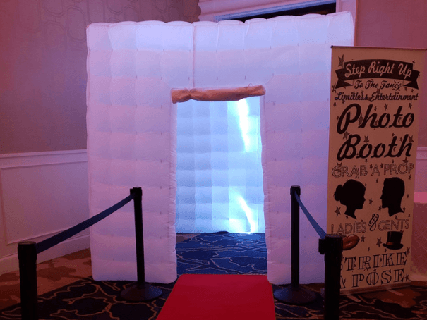 Inflatable photo booth with glowing white walls, a red carpet leading to it. A vintage-style sign invites guests to strike a pose. Festive atmosphere.