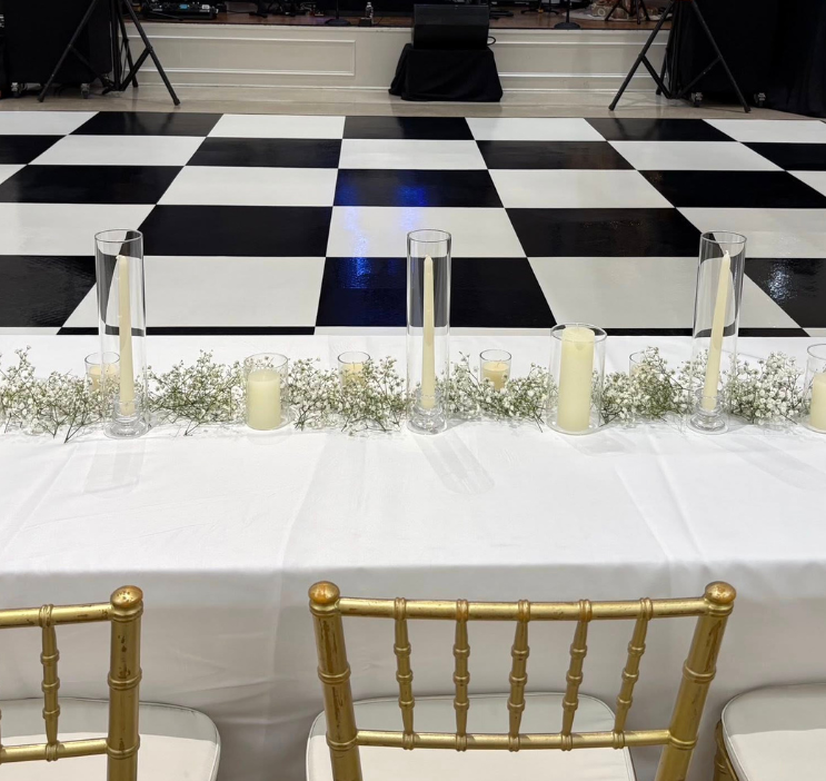 Elegant wedding table with tall glass candle holders and white flowers on a white tablecloth. Gold chairs in front, black and white checkered floor behind.