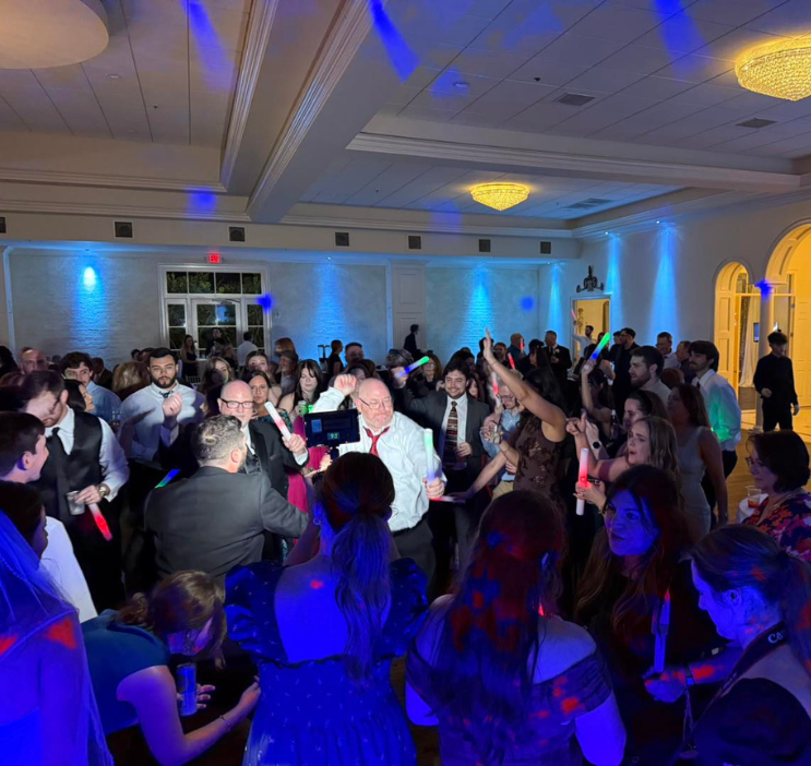 A lively wedding reception with a crowded dance floor, multicolored lights, and guests in formal attire enjoying the celebration under a grand chandelier.