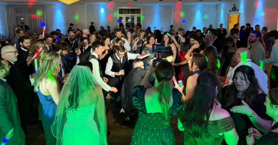 A lively wedding reception dance floor filled with guests in formal attire. Colorful lights reflect off walls, creating a joyful and festive atmosphere.