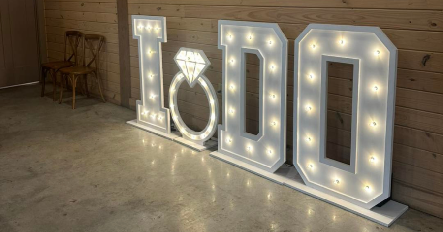 Illuminated letters spelling "I DO" with a ring symbol between them are set against a wooden wall, creating a romantic and celebratory atmosphere.