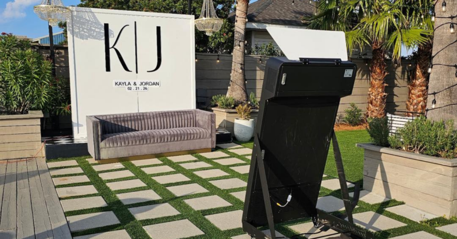 Outdoor event setup featuring a sleek gray couch on a checkerboard grass and stone patio. A backdrop displays "KJ" with date details.