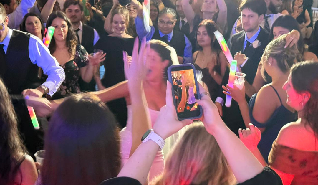 A lively party scene with people dancing energetically, holding glowing sticks. A central figure is captured on a smartphone. The mood is joyful and vibrant. Northshore Wedding Entertainment Ideas