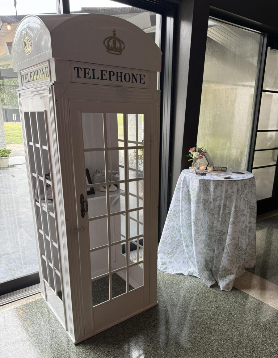 White vintage-style phone booth with crown emblem, next to a round table draped in floral fabric, holding flowers and candles, in a sunlit setting. Audio Guestbooks