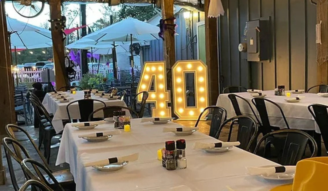 Outdoor party setup with decorated tables, white tablecloths, and empty plates. Large illuminated "40" sign center stage, festive and celebratory ambiance.