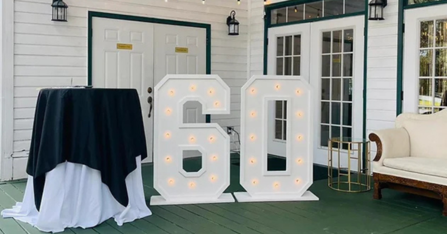 Large illuminated numbers "60" stand prominently on a green wooden deck, surrounded by white walls. Nearby, a round table is draped in black and white, next to a cream sofa, creating a festive atmosphere.