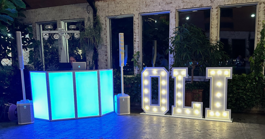 DJ booth with glowing blue panels and a laptop setup, flanked by speakers. Large illuminated letters spell "Oli" against a backdrop of plants and windows.