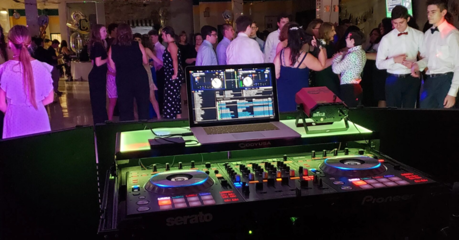 A DJ booth with colorful lights and turntables is in the foreground, while people in formal attire dance and socialize in the background at a lively party.