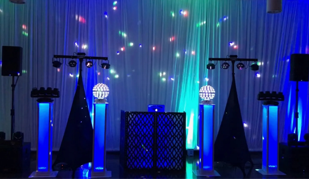 A vibrant DJ setup with glowing blue columns, disco balls, and colorful LED lights against a backdrop of white curtains, creating a lively party atmosphere.